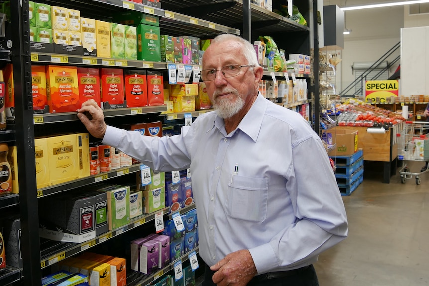 An elderly man with a blue button up shirt stands next to the grocery shelves in a retail store