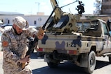 A member of Misrata forces wearing army clothing points and prepares a gun with an army truck in the background.