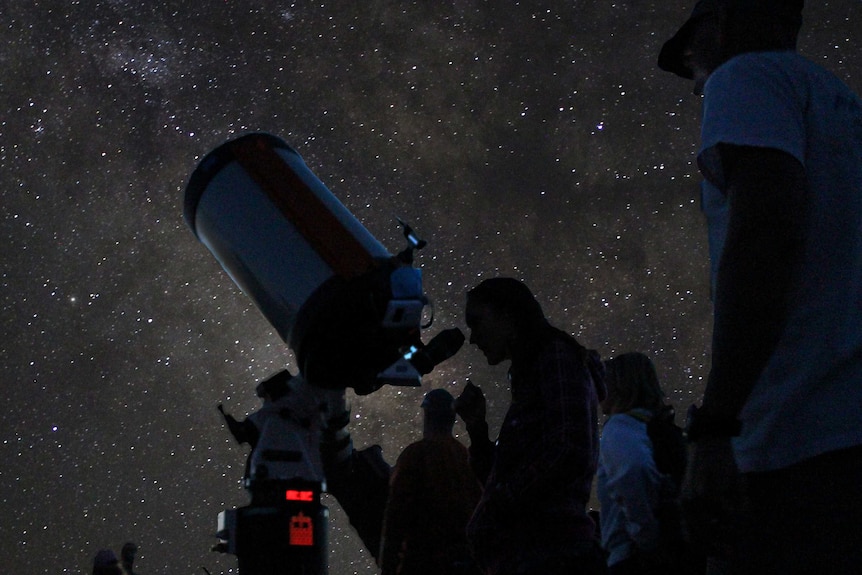 A starry night sky in the background with several silhouetted people looking through telescopes.