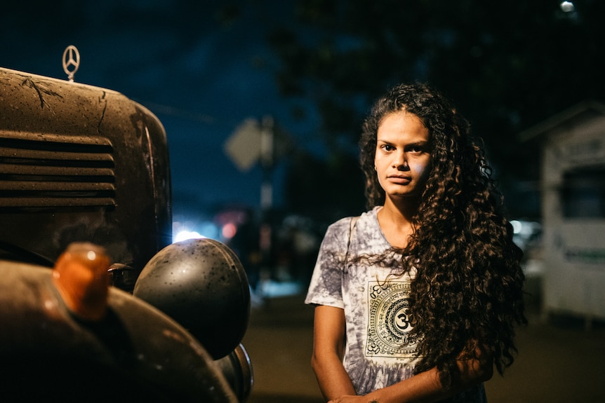 A woman with long brown curly hair sits next to an old car.