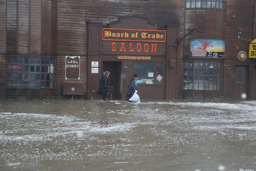 two people walk down a flooded street past a brown building marked saloon