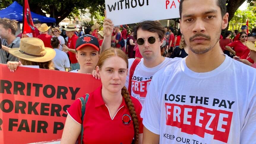 A group of people wearing red shirts protesting in a park holding signs and looking unhappy