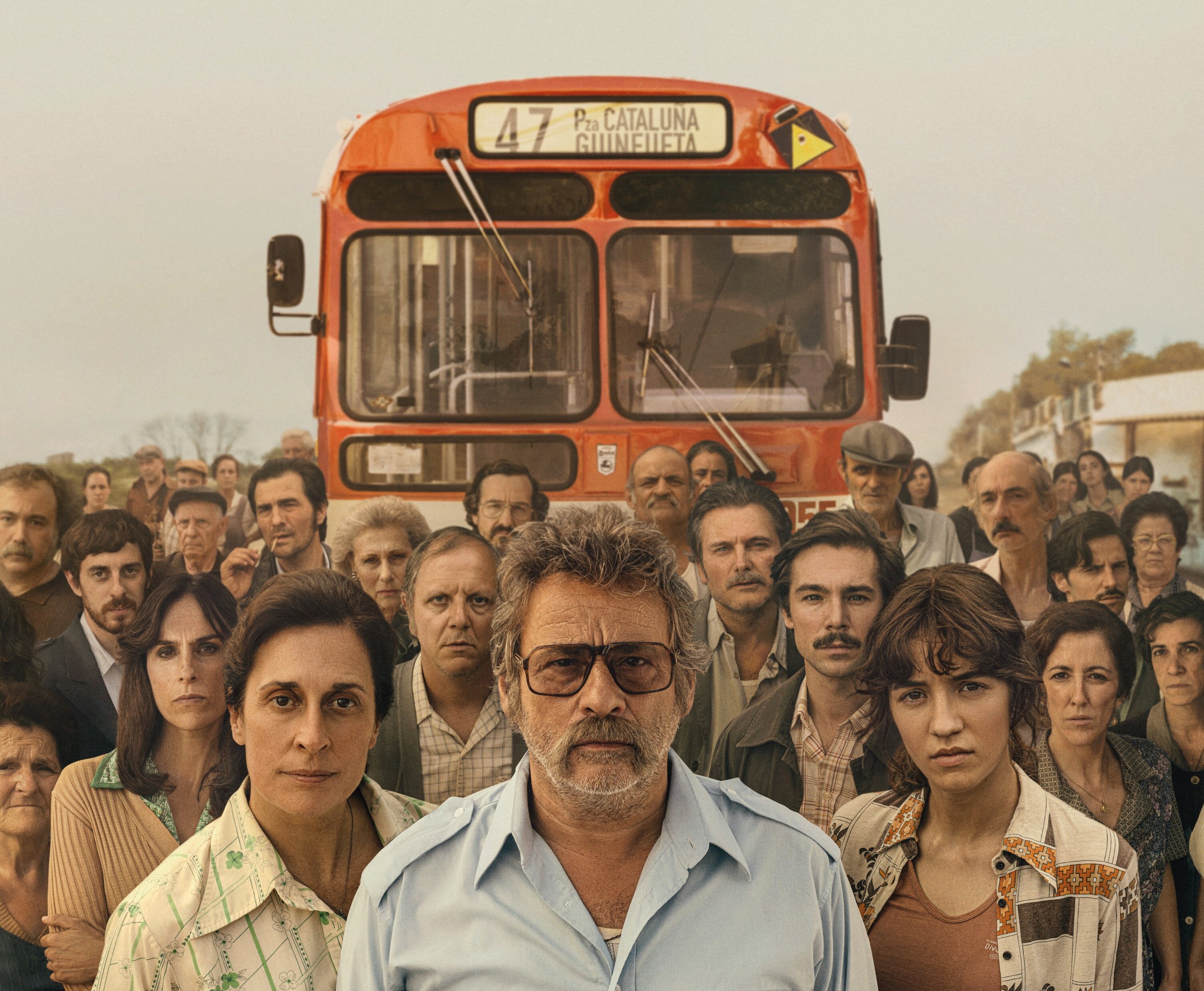 A man wearing a 1970s bus driver uniform and sunglasses stands in front of a large group of people and a bus.