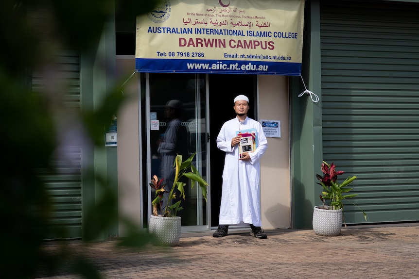 A man holding school books stands in front of a sign reading 'Australian International Islamic College'