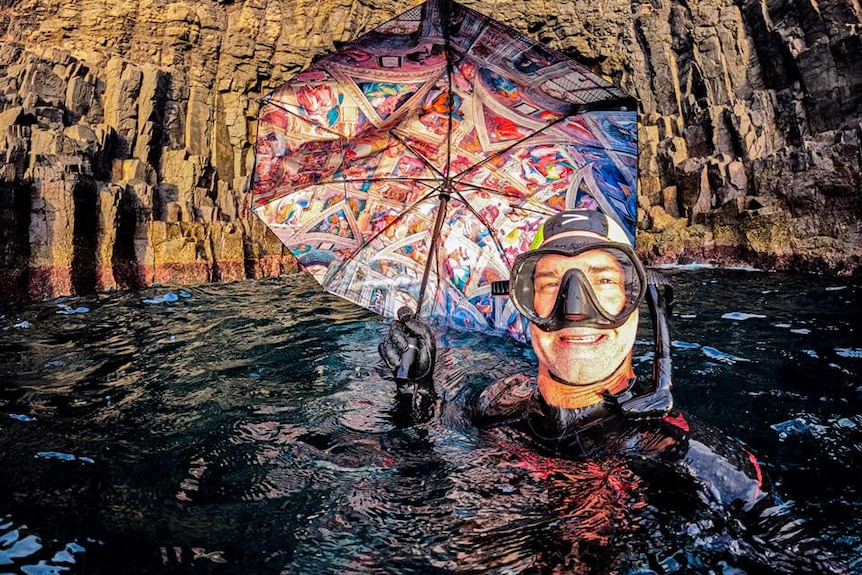 Scott Gutterson holds a colourful umbrella while wearing snorkelling gear and submerged in the water. 