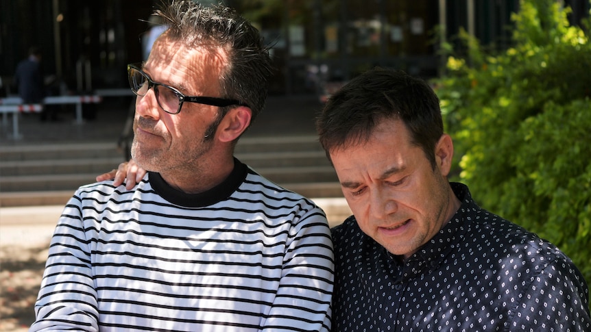 A man with glasses in a stripe shirt and another man in a dark blue shirt with his arm around his shoulder