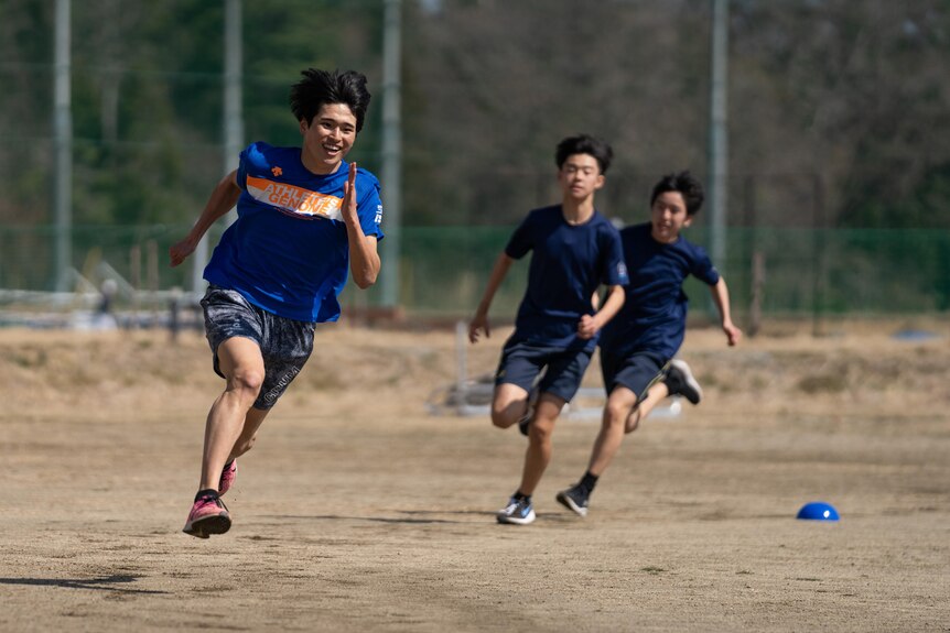 A Japanese boy grinning and running while two boys run at a distance behind him. 
