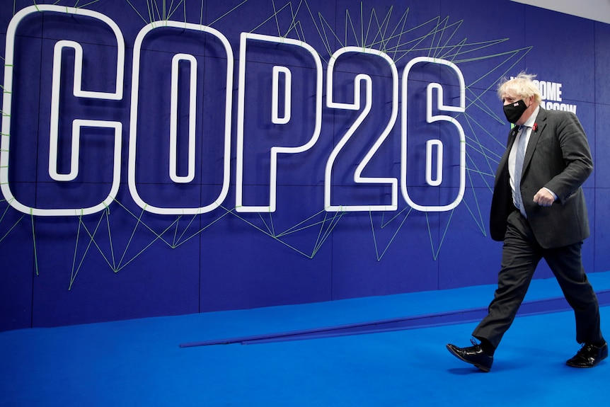Man in a black suit wearing a blue tie with white hair walking behind a blue wall with the words 'COP26.' 