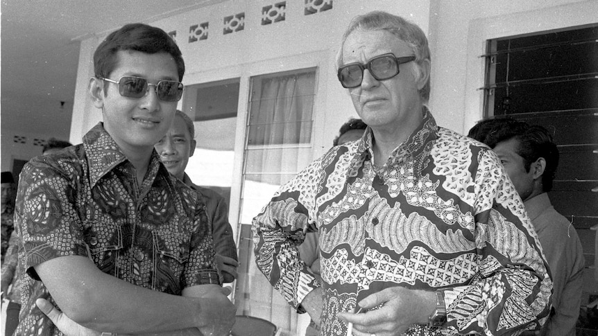 black and white photo of an older white man wearing a batik Indonesian shirt and dark sunglasses with a younger Indonesian man