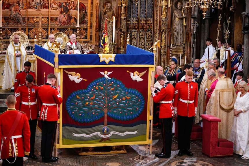 Guards stand around a screen with a patchwork tree and angles on it. 