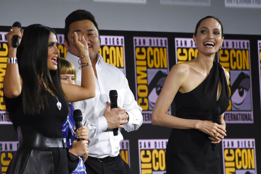 Salma Hayek, Lia McHugh, Don Lee and Angelina Jolie laugh together on stage, holding microphones.