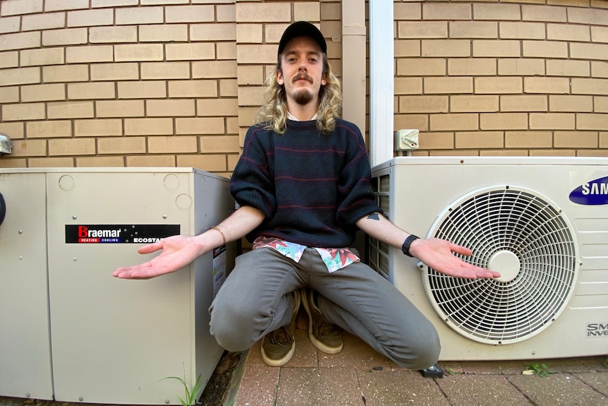 A man crouches between two AC outdoor units.