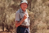 An old man wearing a hat and holding a divining rod standing in bushland.