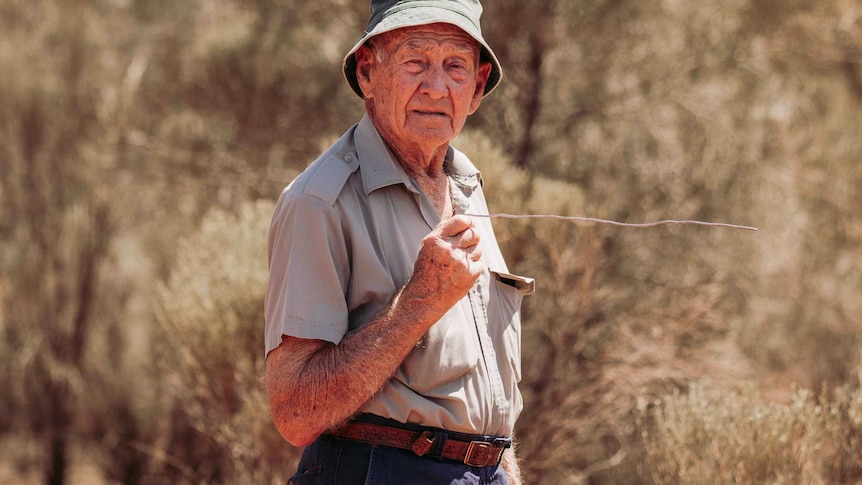 An old man wearing a hat and holding a divining rod standing in bushland.