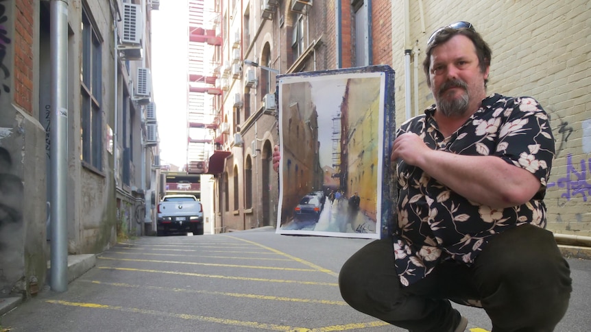 A man holding a painting of an alley, while standing in the same alley, looking to camera.