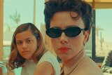 A woman of colour in a white dress and a white woman with brunette 50s-styled curls, red lipstick and sunglasses sit in a diner.