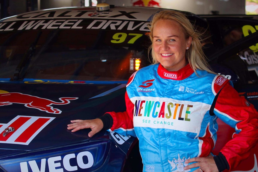 A woman wearing a Supercars drivers' suit with a Newcastle logo leans on a racing car.