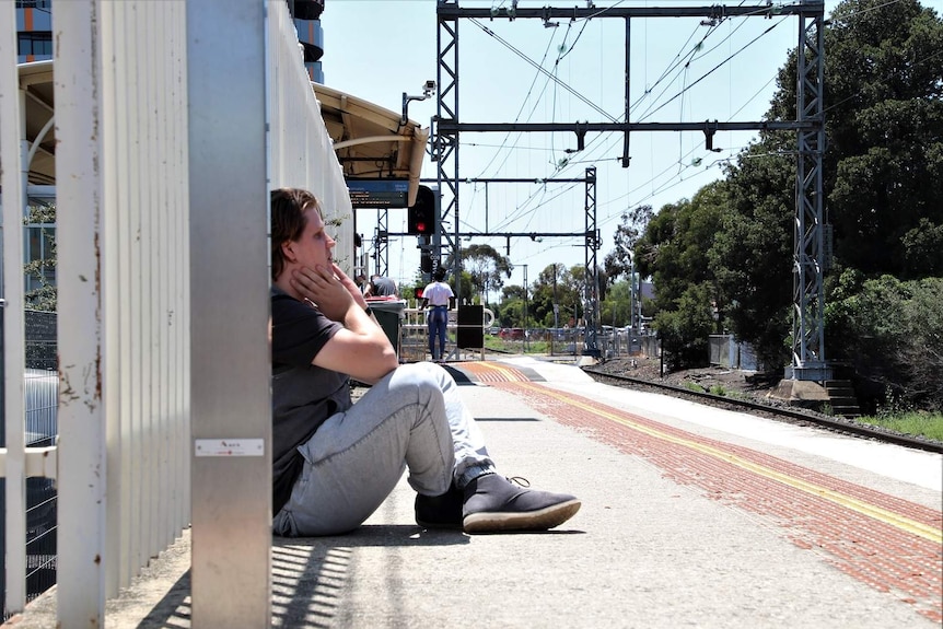 Allan sits on a train station platform looking worried.