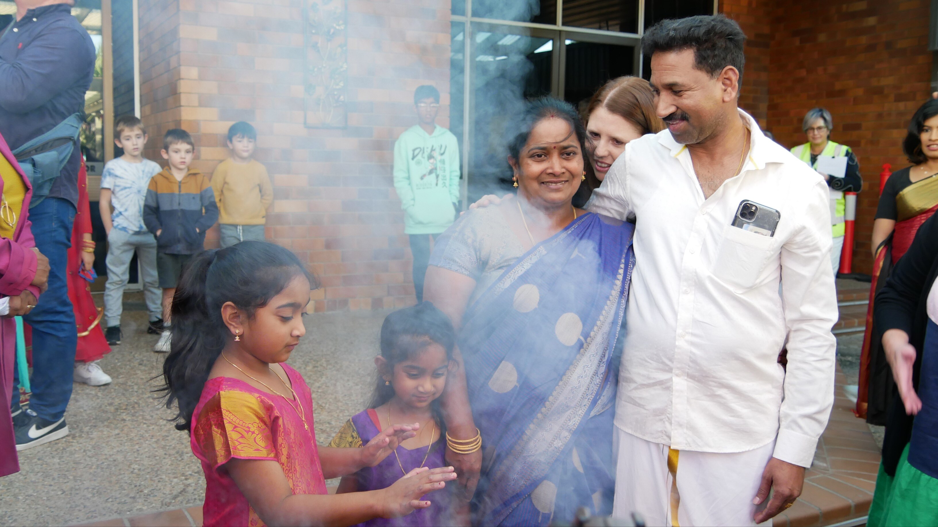 Two young girls and their parents, in traditional Tamil outfits, stand behind some smoke