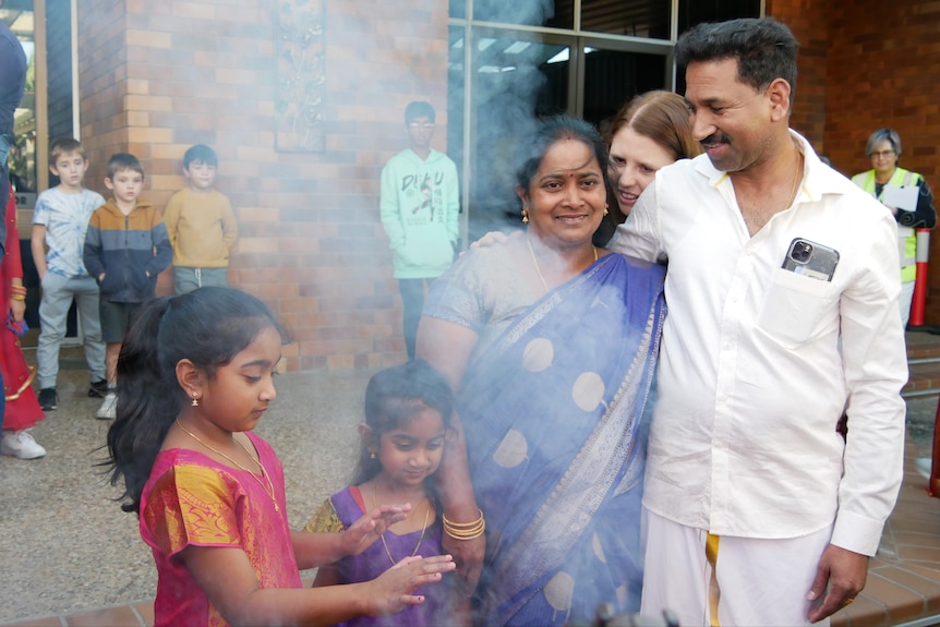 Two young girls and their parents, in traditional Tamil outfits, stand behind some smoke
