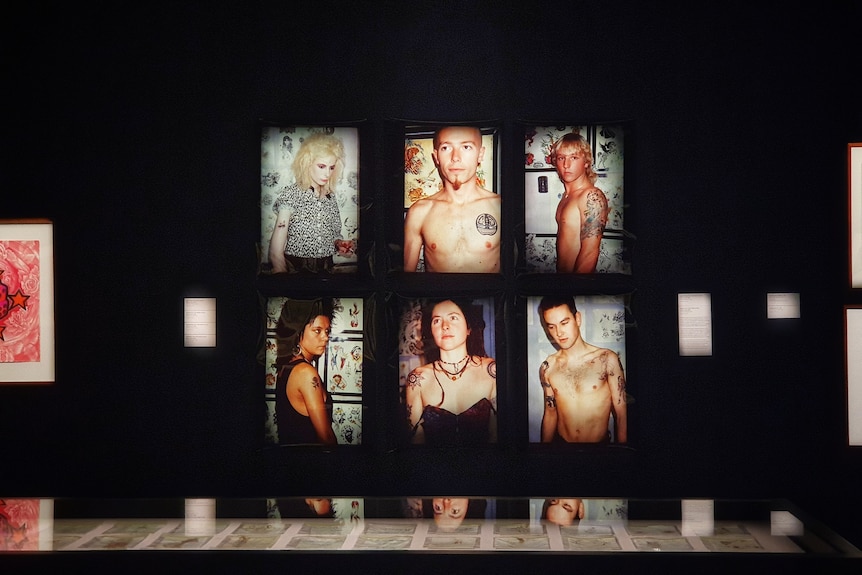 Six portraits of people showing off their new tattoos hang on  a  black gallery wall.