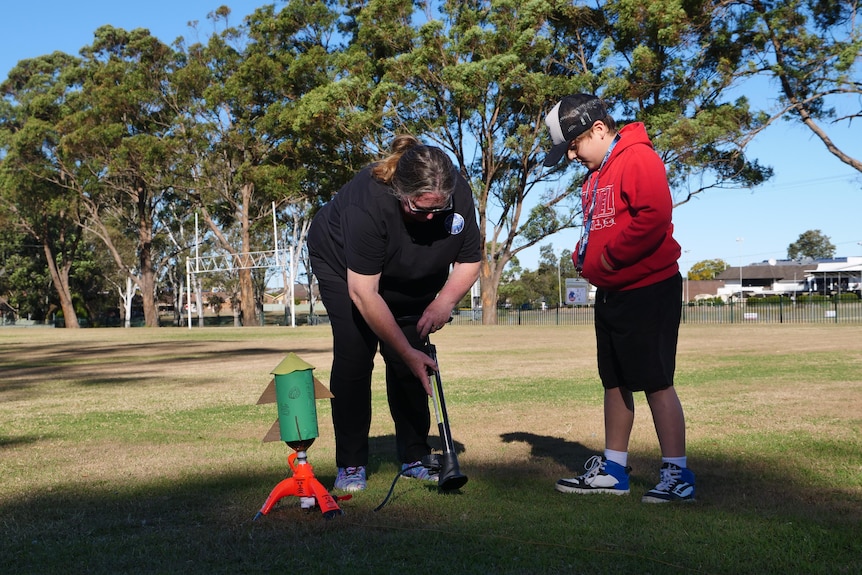 Woman in black with a child in red jumped helping to pump up a green rocket model.