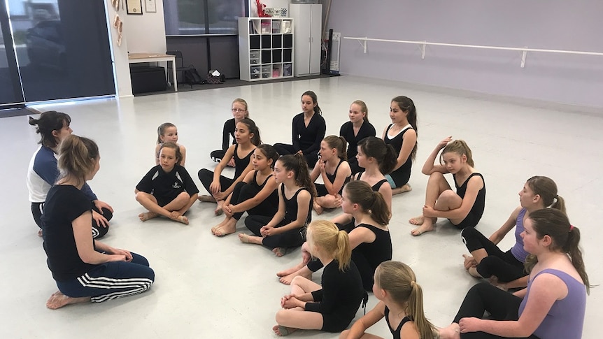 Dance students sit on the floor listening to two dance instructors