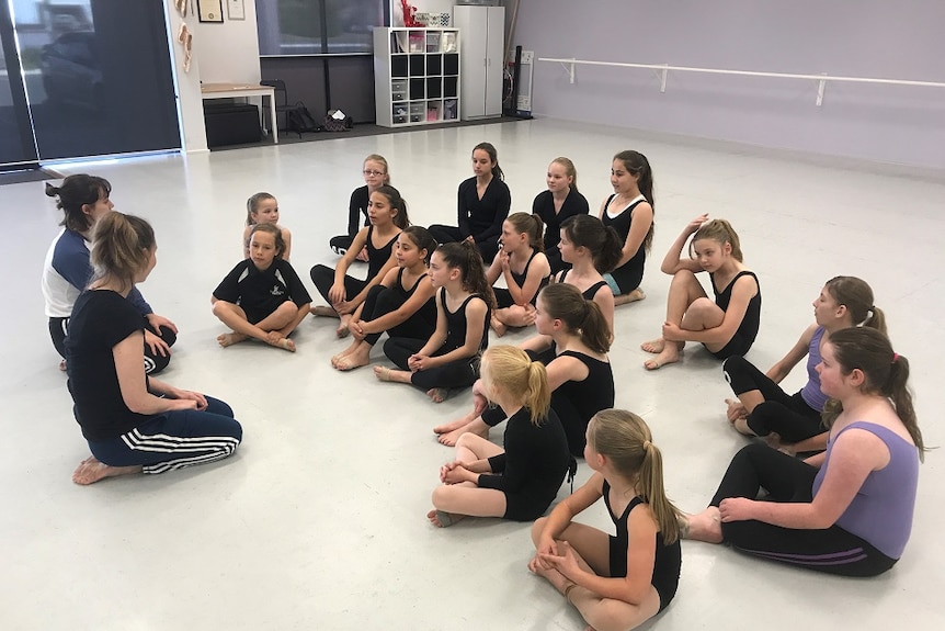 Dance students sit on the floor listening to two dance instructors