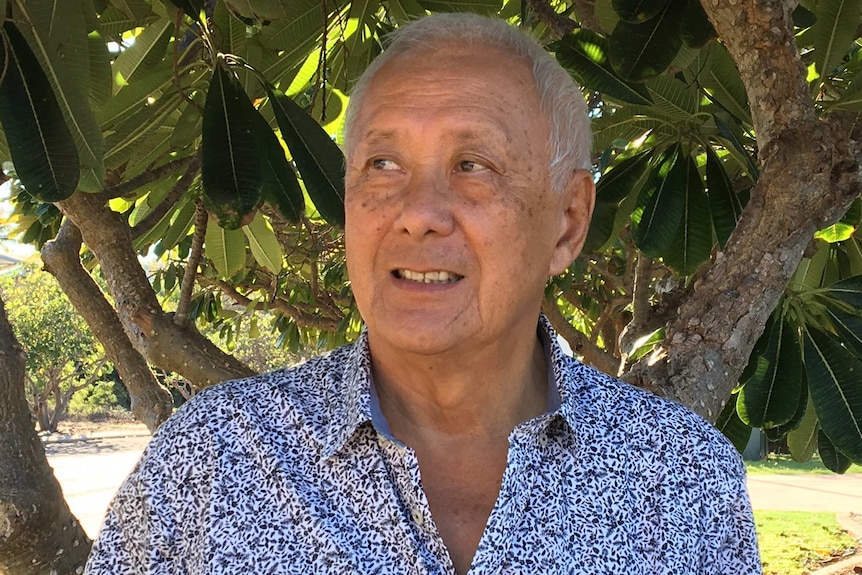 A man in a blue and white shirt smiles and looks to the side of the camera as he stands in front of a tree.