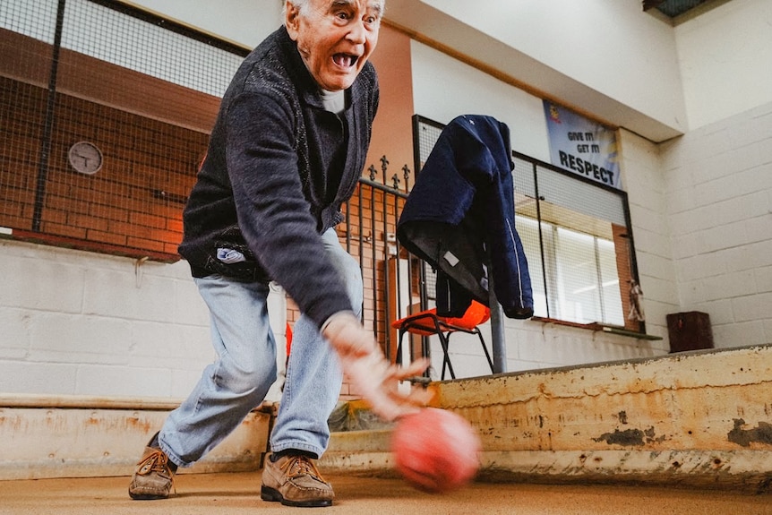 An old man smiles as he bowls a bocce ball down a sand lane indoors.