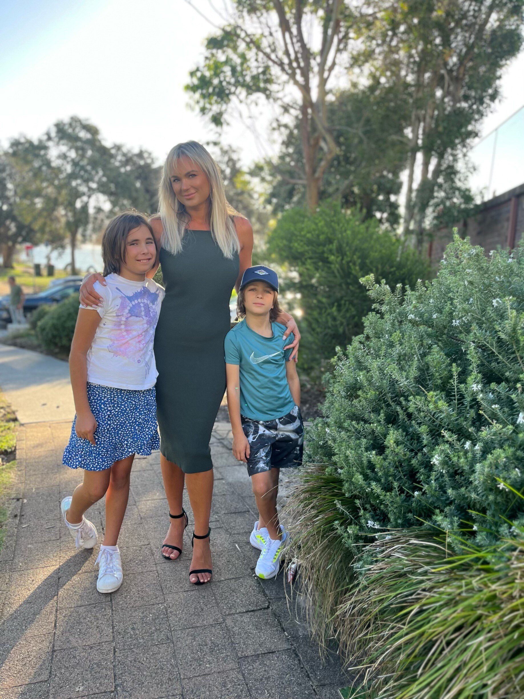 A mum poses with two kids on the sidewalk outside a house.