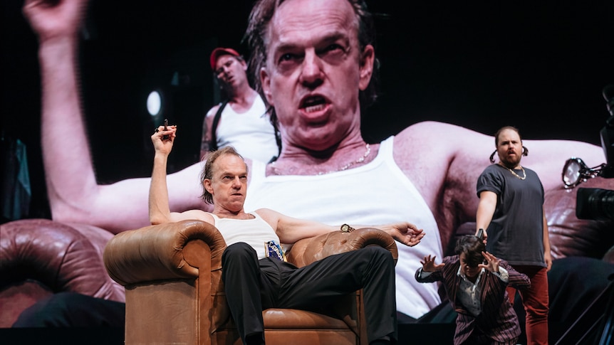 Actor Hugo Weaving on stage sitting in armchair wearing white tank singlet and black suit pants and shoes.