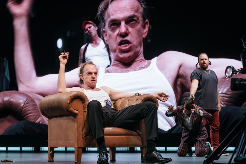 Actor Hugo Weaving on stage sitting in armchair wearing white tank singlet and black suit pants and shoes.