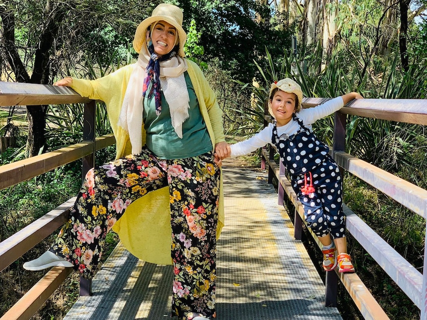Aseel and her daughter hold hands and smile on a bridge, wearing colourful clothes.