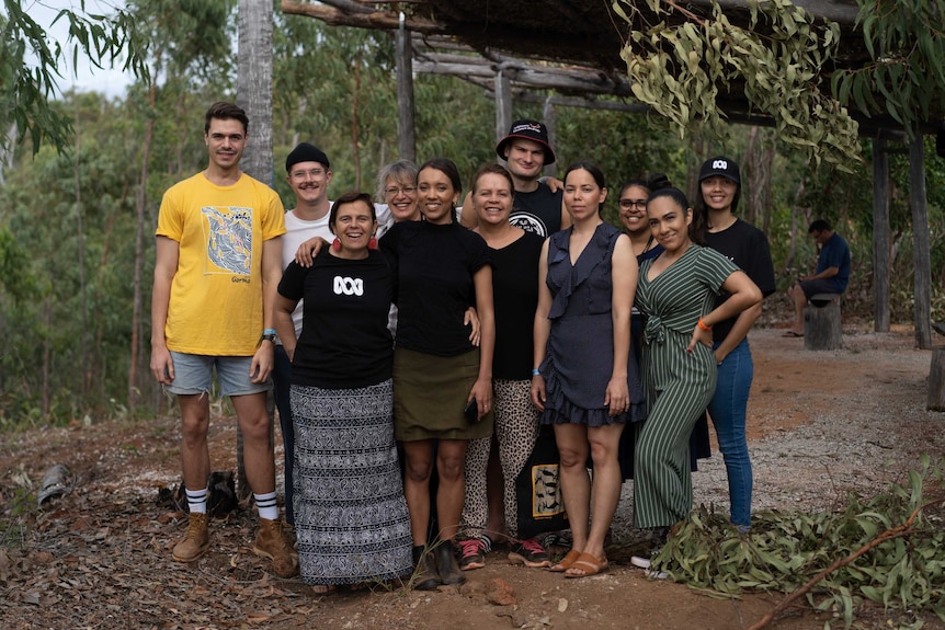 Eleven people standing with arms around each other, some wearing ABC t-shirts or caps, with a bush background.