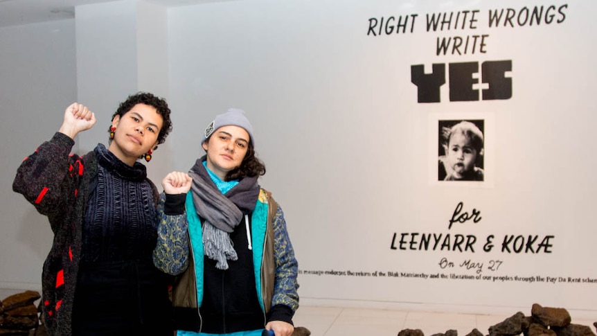Two young women hold up fists in a white room, rock formation on floor, text on back wall reads 'right white wrongs, write yes'.