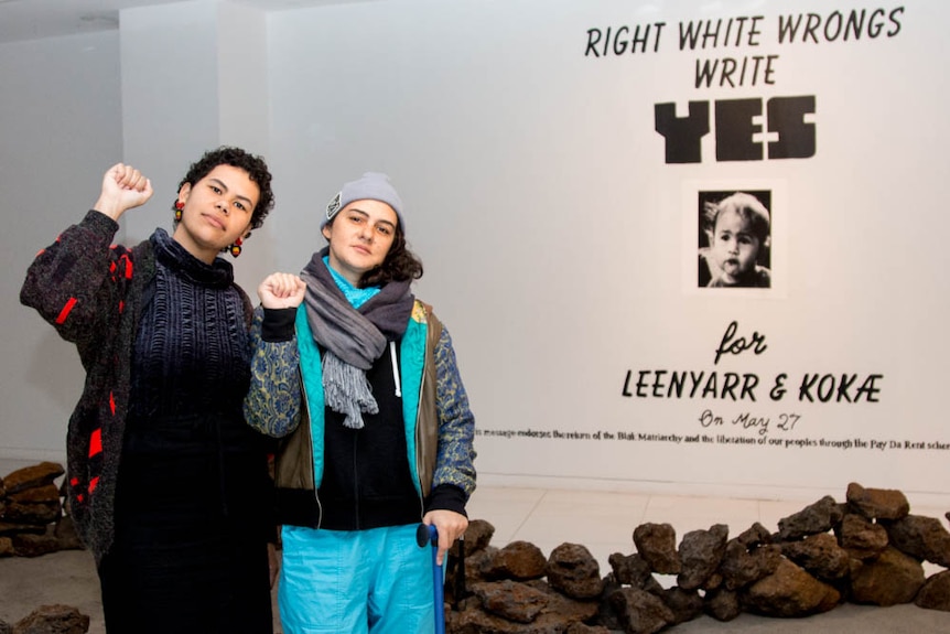 Two young women hold up fists in a white room, rock formation on floor, text on back wall reads 'right white wrongs, write yes'.