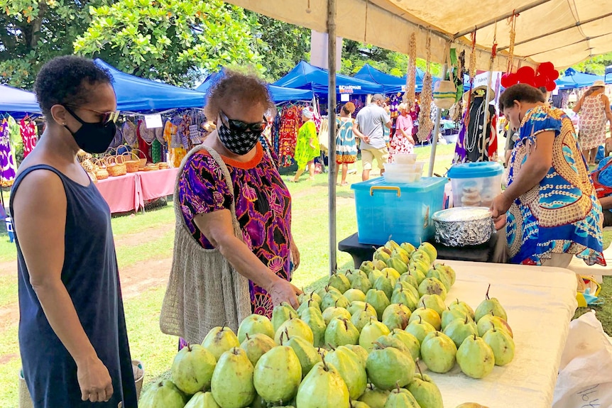 Two women in face masks inspect a table stacked with pears at a market