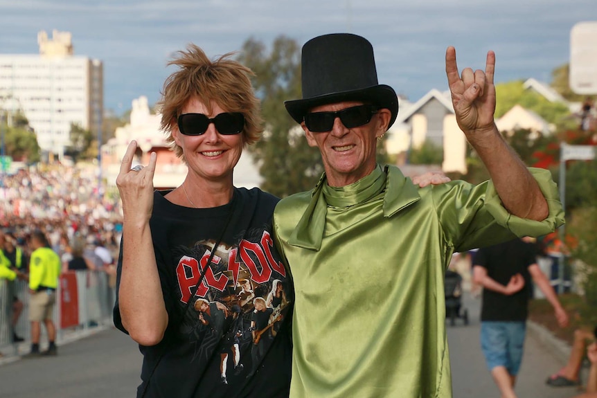 Two people, one wearing an AC/DC shirt and the other a green top and top hat, pose for a photo at a festival showing rock horns.