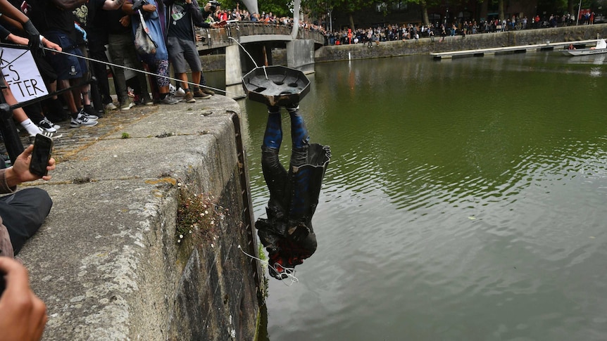 A crowd lowers a statue into a harbour