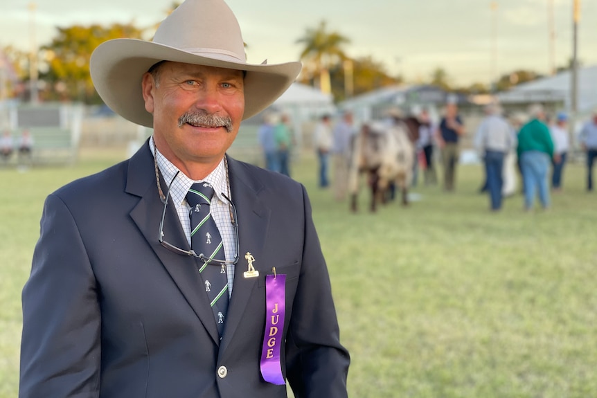 A man with a grey moustache, suit and large hat standing in front of a blurred background.