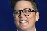 Hannah Gadsby stands in front of a microphone wearing a navy suit and holding a presenter's information card