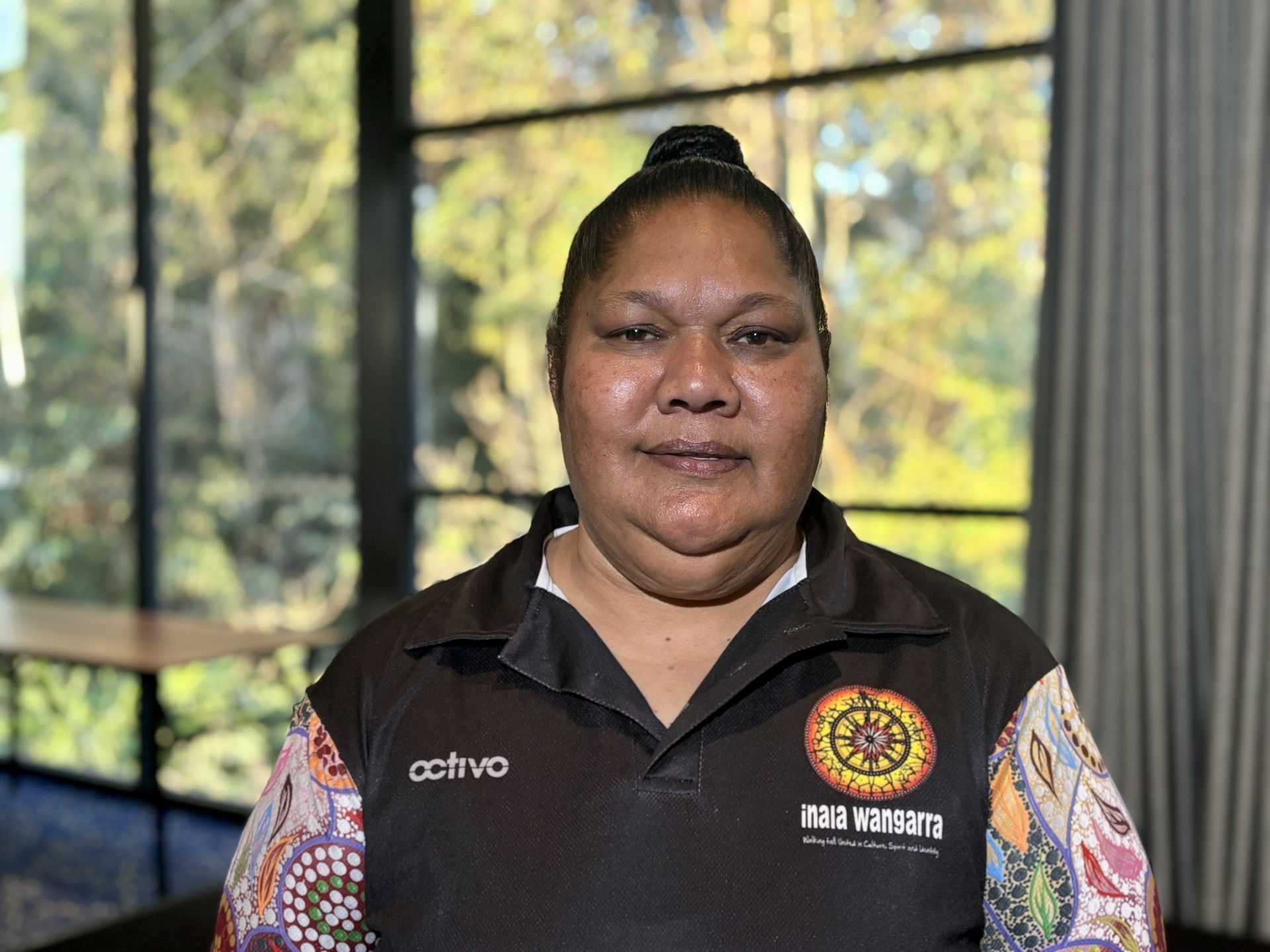 a woman looks down the barrel of a camera while wearing an indigenous-themed black polo shirt