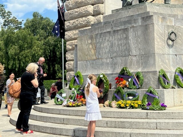 Two people look up at a war memorial that has wreathes laid at the base.