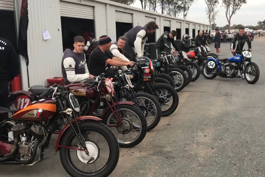 A group of motorcycle riders at a race day sit atop their vintage indian motorcycles.