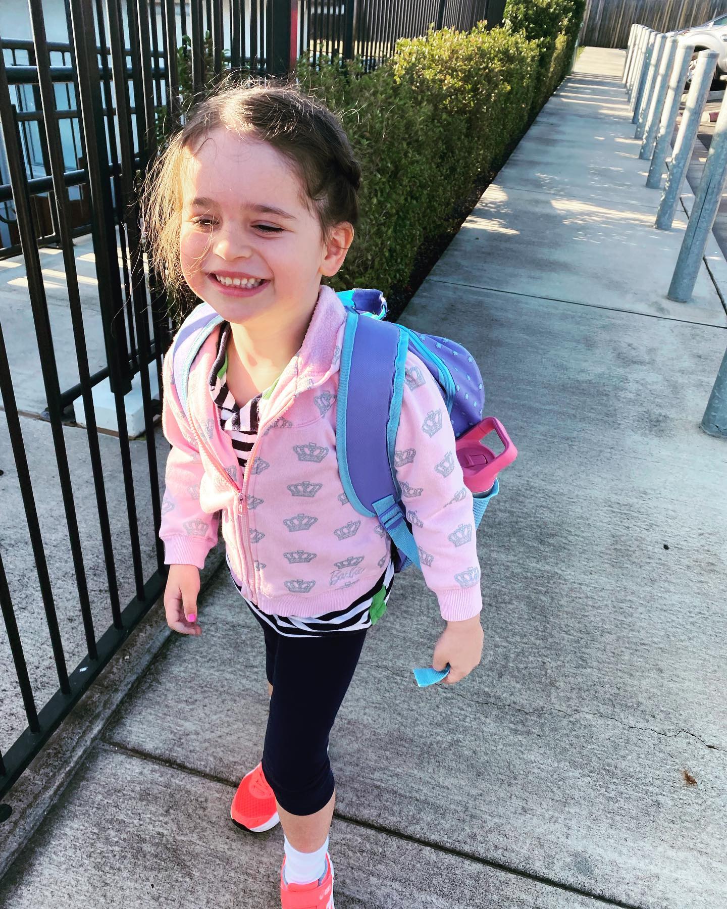 Young girl, Arlo, smiling, walking, wearing a pink jumper and purple backpack.