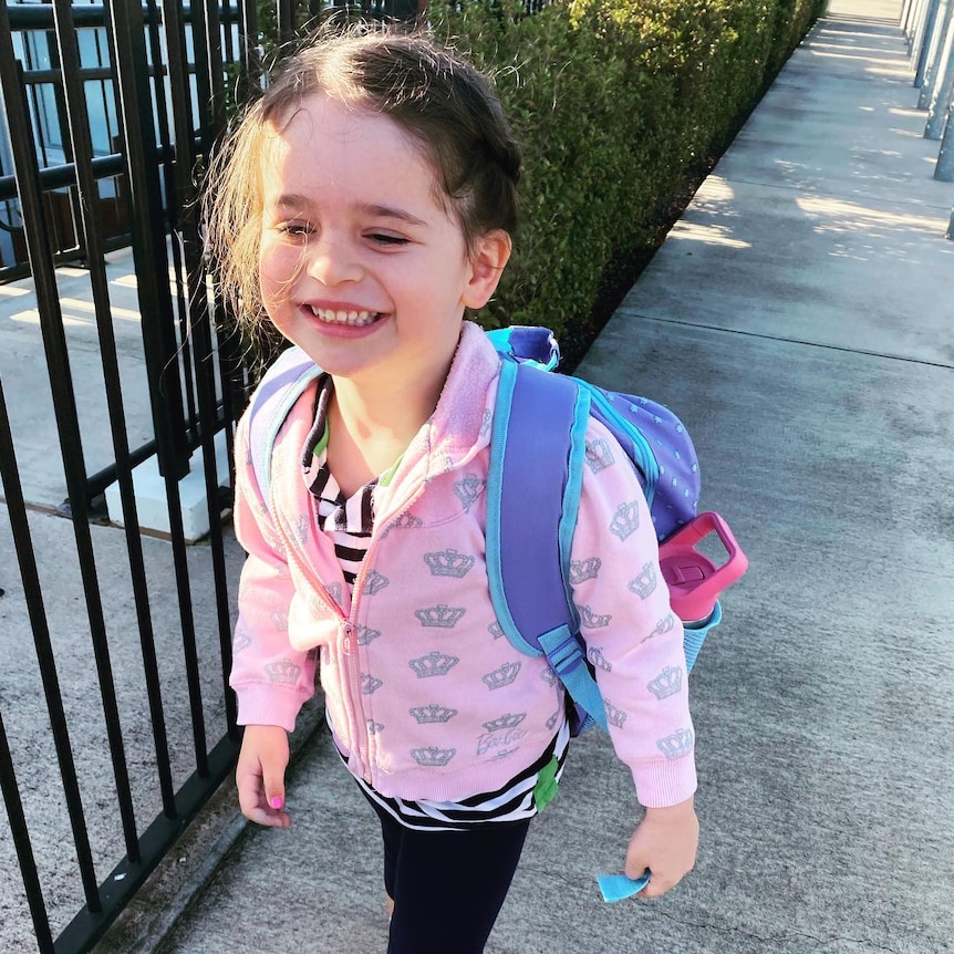 Young girl, Arlo, smiling, walking, wearing a pink jumper and purple backpack.