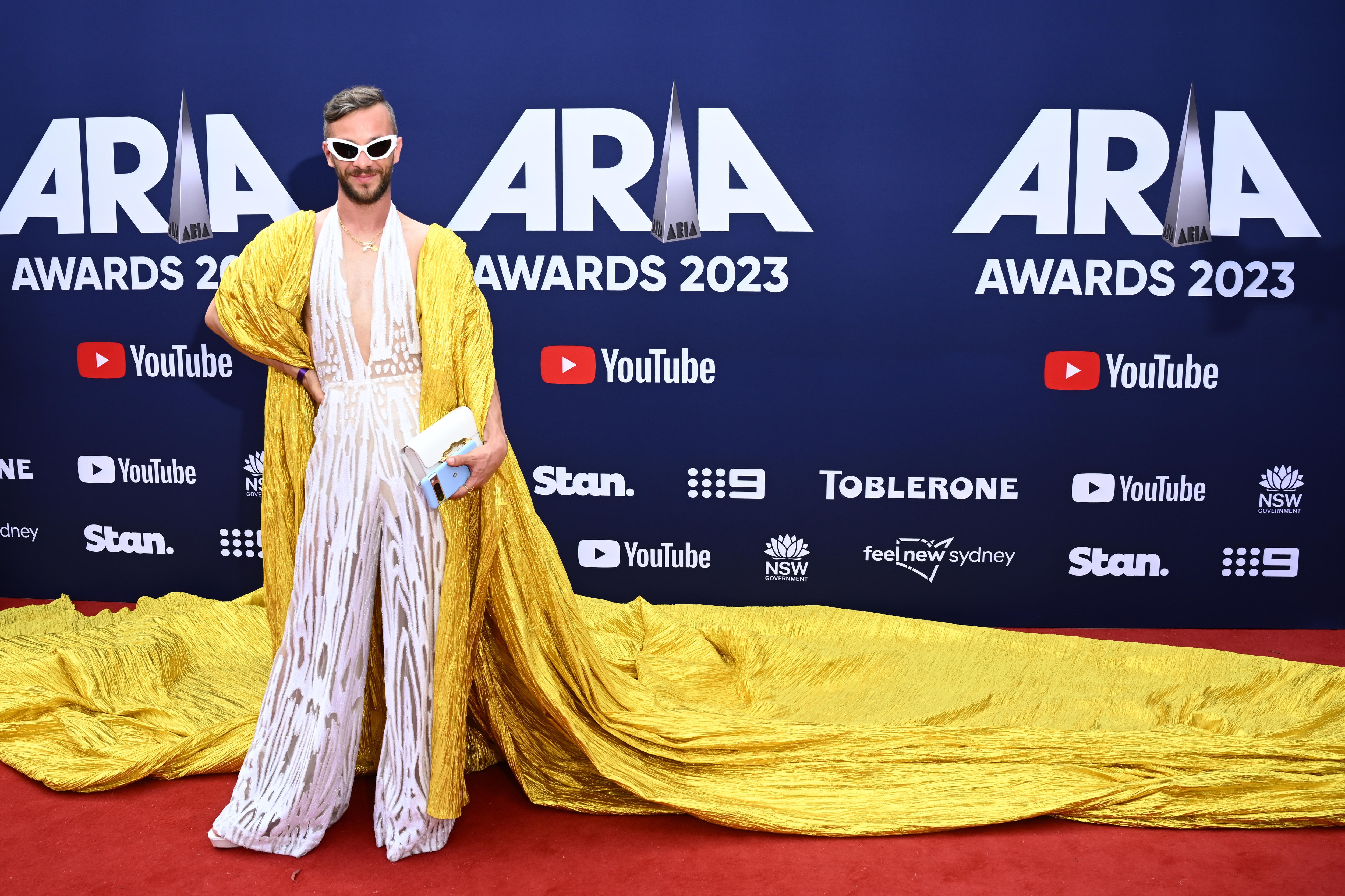 A man wearing a white playsuit with a yellow cape and sunglasses