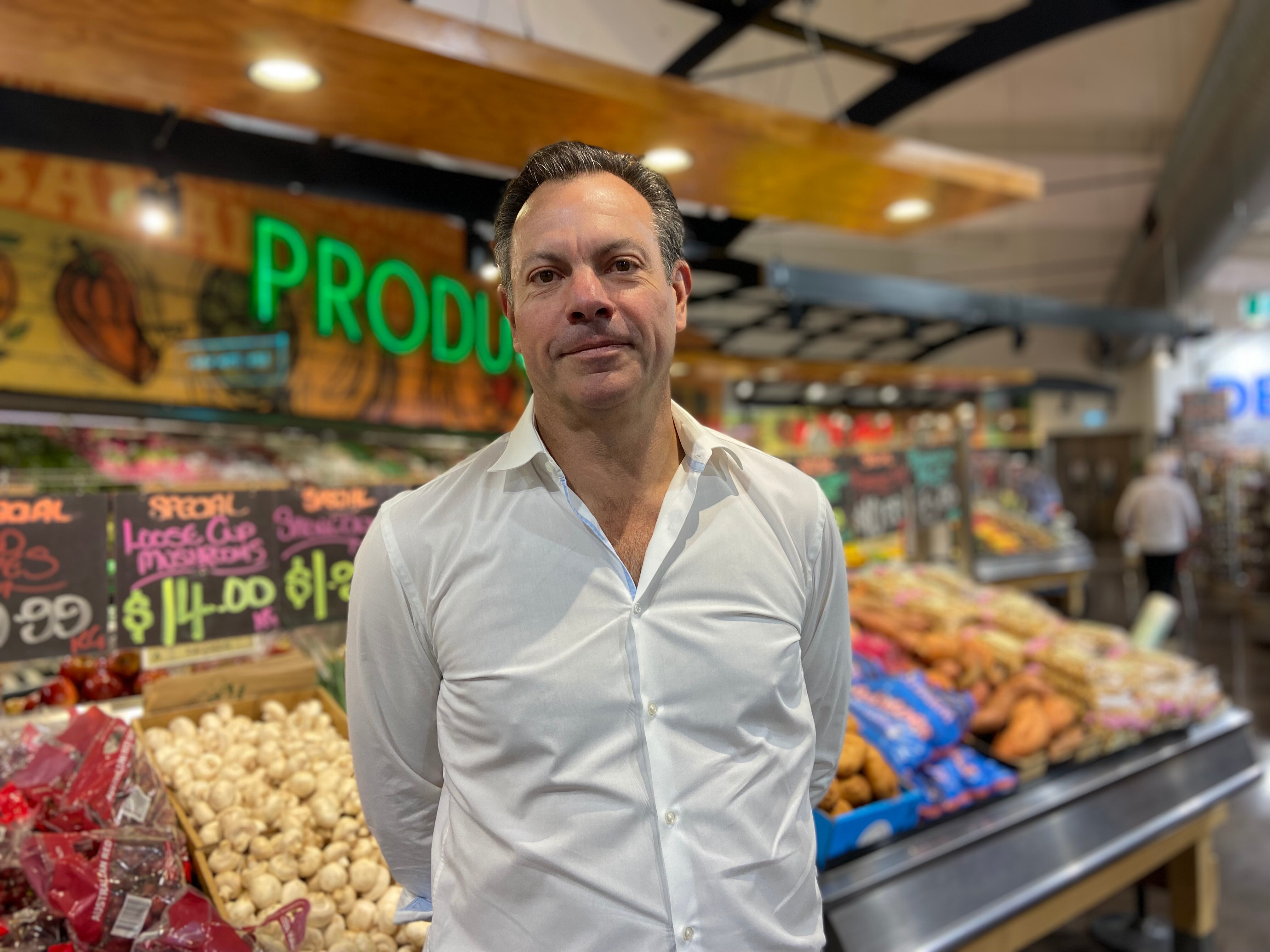 a photo of a man in white shirt standing in front of vegetables in supermarket 