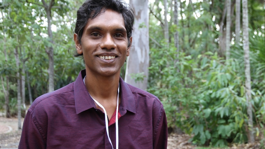 A young man stands on a footpath and smiles at the camera.